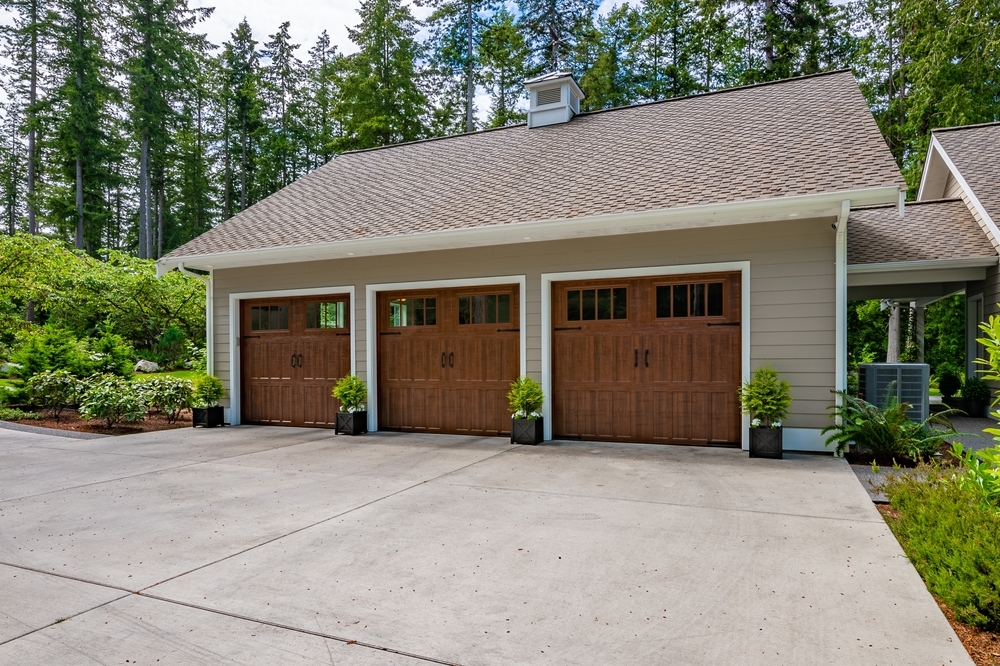 A three-car garage with wooden doors, surrounded by greenery and trees, with a concrete driveway in front.