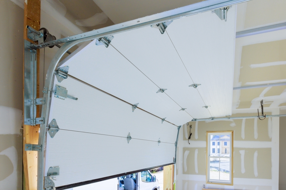 Inside view of a partially open garage door with visible metal hinges and a mechanism track. Walls are unfinished with drywall and a small window on the right.