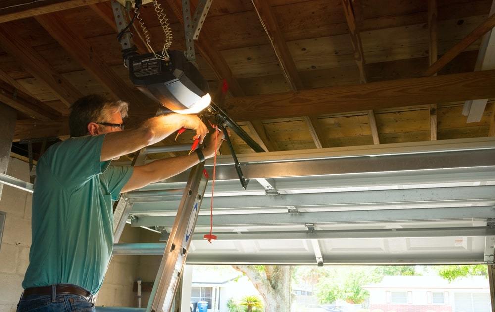 A man on a ladder adjusts a garage door opener inside a garage with an open ceiling.