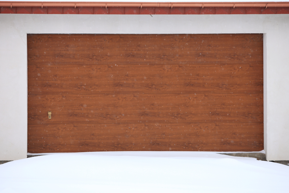 A closed brown wooden garage door with a layer of snow on the ground in front.