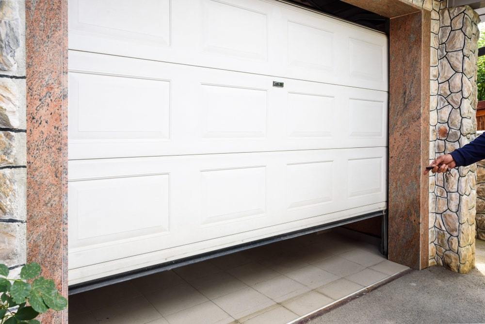 A partially open white garage door with a remote control in someone's hand. The surroundings include stone walls and a tiled floor.