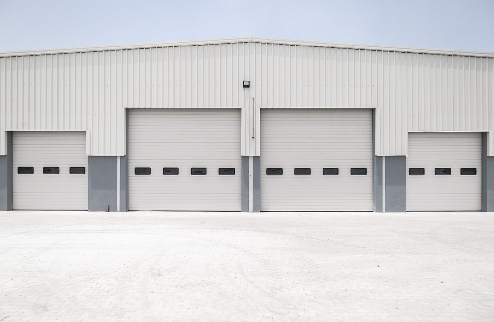 A large warehouse with three closed, white roll-up doors. The building has a gray lower section and a smooth, light concrete foreground.