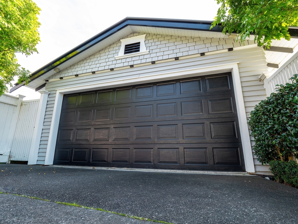 A black garage door of a white house with a shingled facade and surrounding greenery.