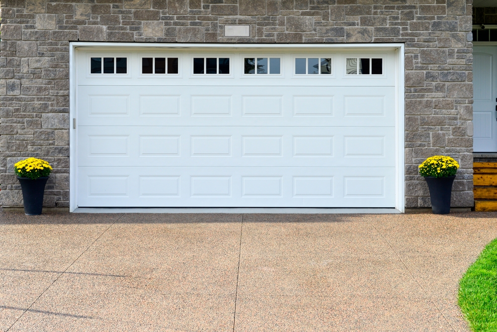A closed white garage door with rectangular windows at the top, flanked by potted yellow flowers, set in a stone wall with a concrete driveway in front.