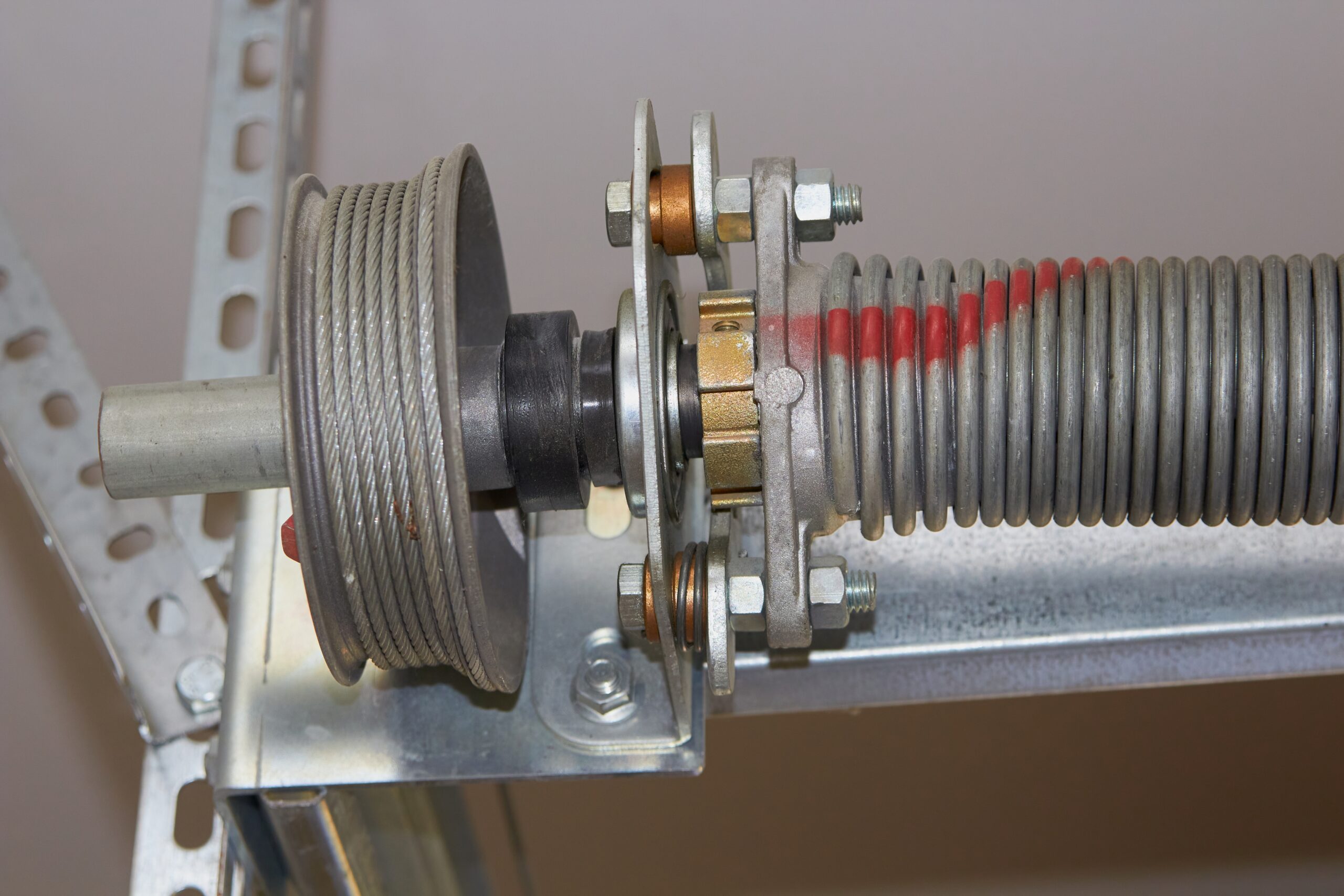 Close-up view of a garage door torsion spring assembly, showing the cable drum, spring, metal bracket, mounting hardware, and a broken garage door cable.