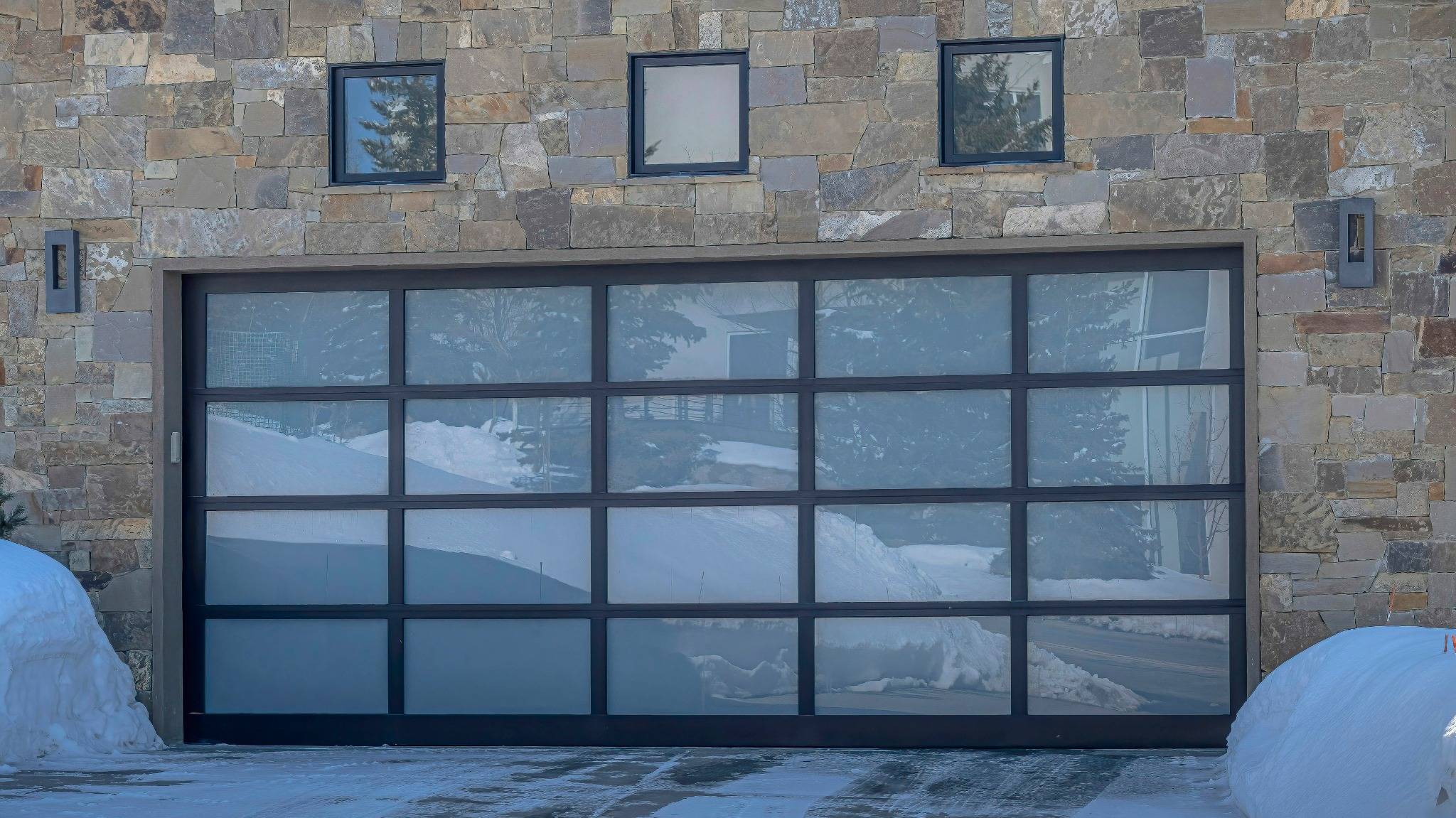 Modern glass-paneled garage door with reflections of snow and trees, set in a stone wall with three square windows above.
