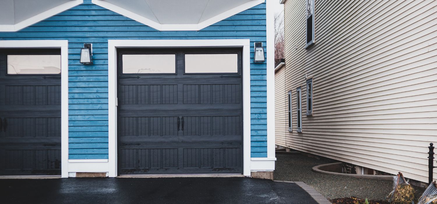 A blue garage with a black door, plus two wall-mounted lights next to a beige-sided house, separated by a narrow gravel path.