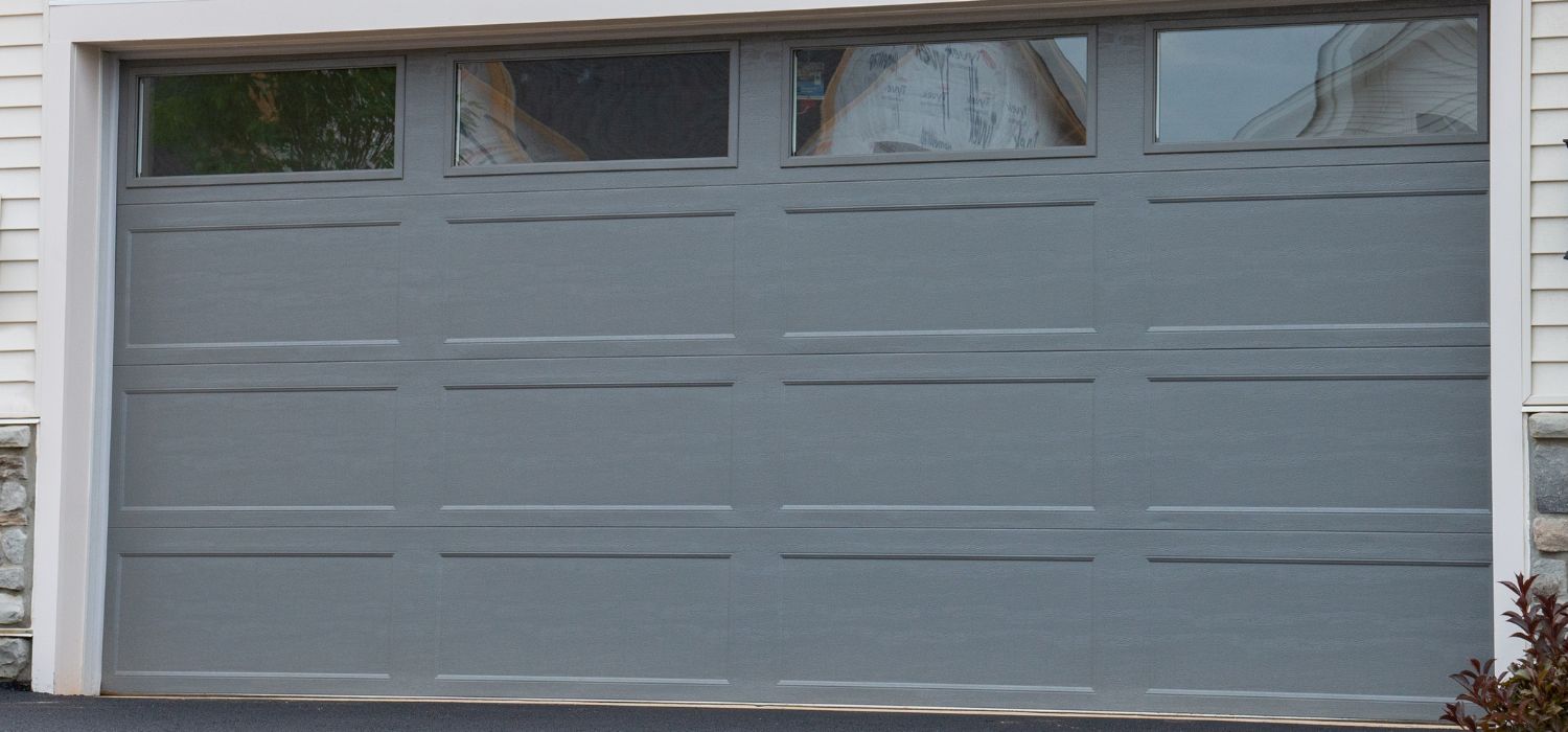 Closed gray modern garage door with four rectangular windows along the top, set in a white exterior wall with stone accents at the edges—an elegant choice inspired by modern garage doors Pinehurst homeowners prefer.