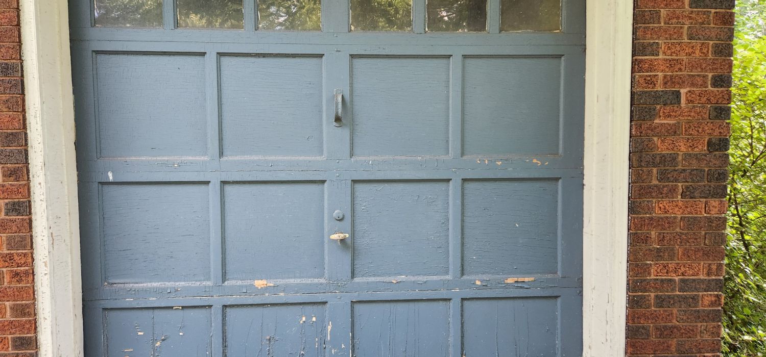 A weathered blue wooden garage door with peeling paint, set in a red brick wall with white trim, and bushes visible on the right side.